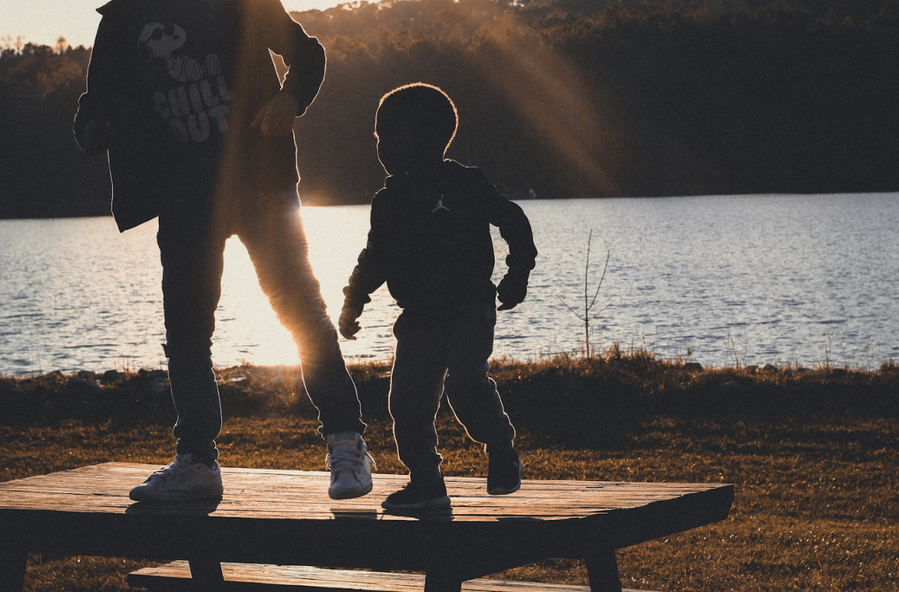 兩個兒童（或較年長者和幼童）站在湖邊的木製平台上，背景是湖水與日落，象徵童年與關係